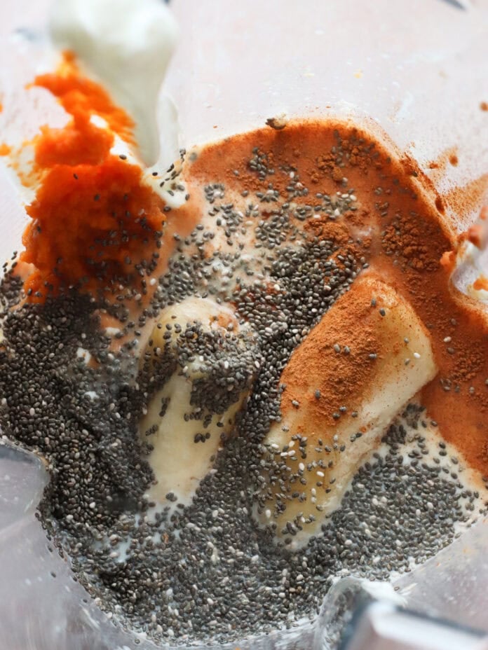 Close-up of a blender containing chia seeds, banana, pumpkin puree, cinnamon, and yogurt, ready to be blended.