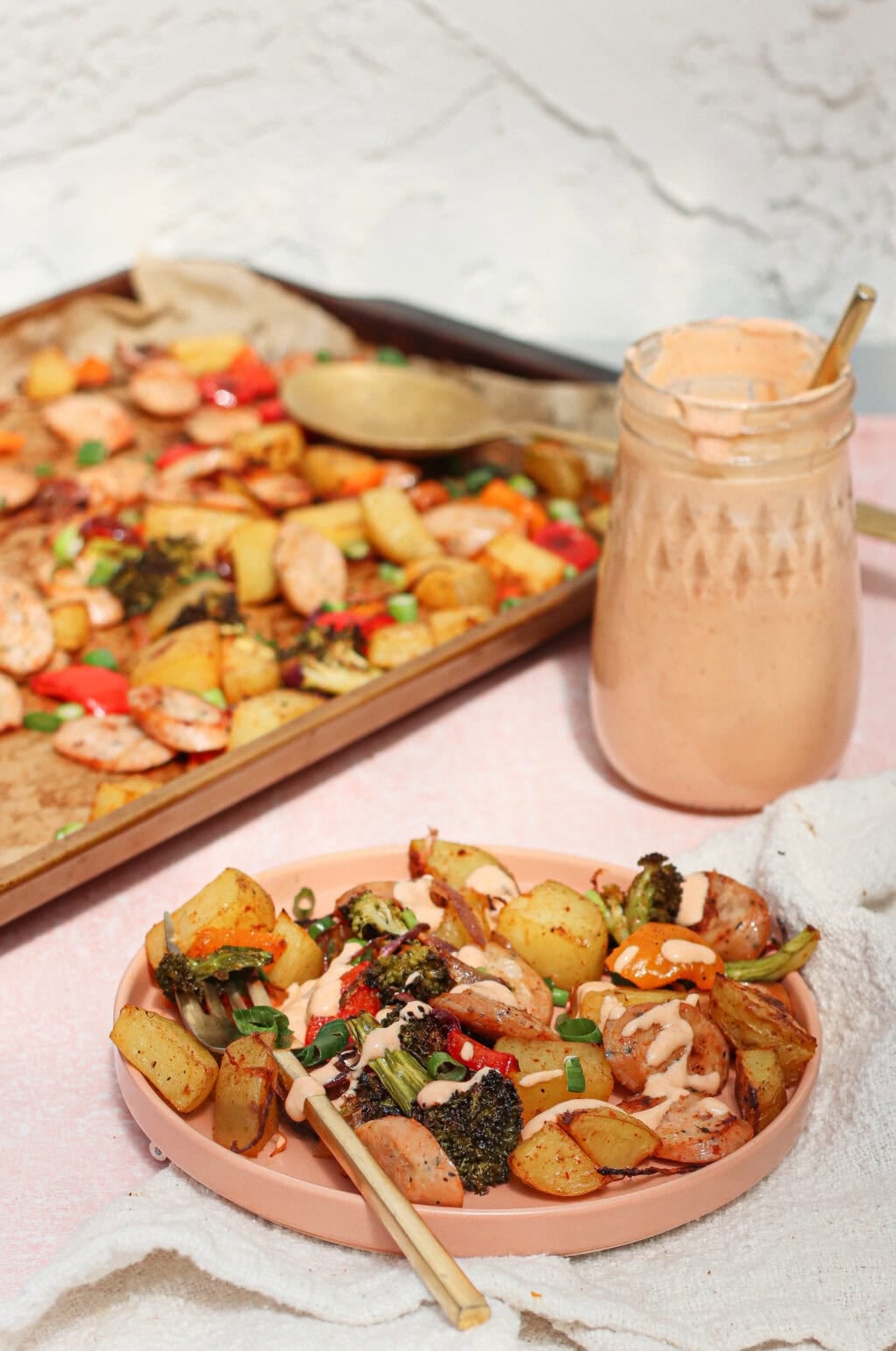 A plate of roasted potatoes, broccoli, and sausage drizzled with a creamy sauce, with a baking tray of the same dish and a jar of sauce in the background. Chopsticks rest on the plate.