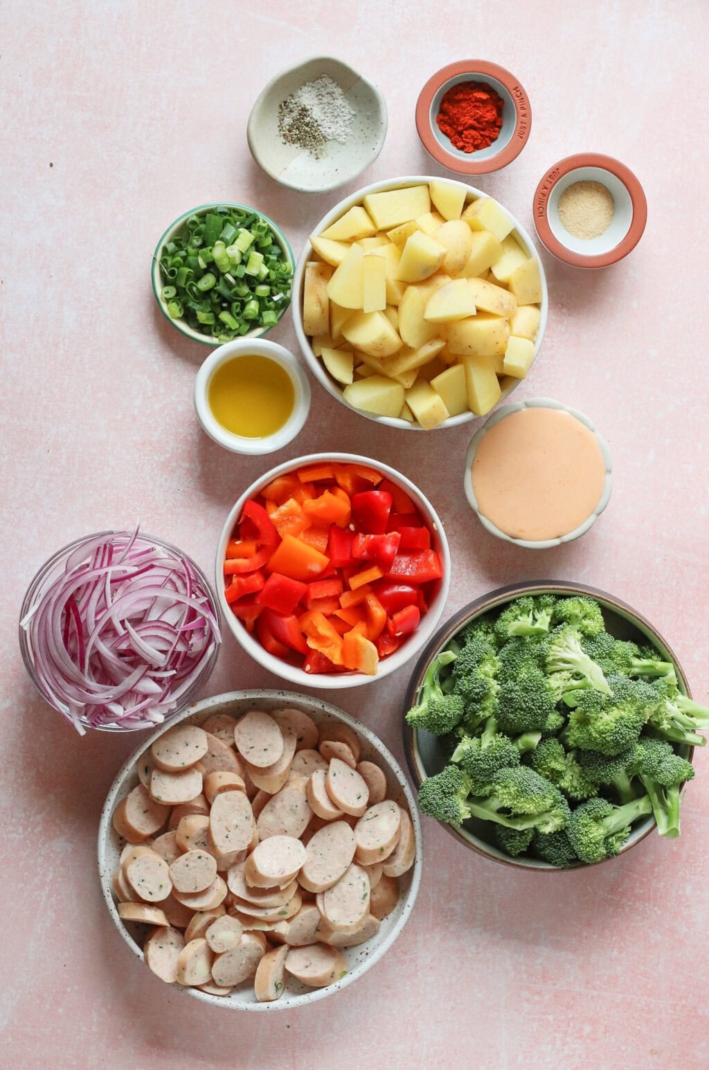 An overhead view of bowls filled with chopped potatoes, broccoli, sliced sausage, red onion, diced red peppers, green onions, olive oil, spices, and a creamy sauce, all arranged on a pink surface.
