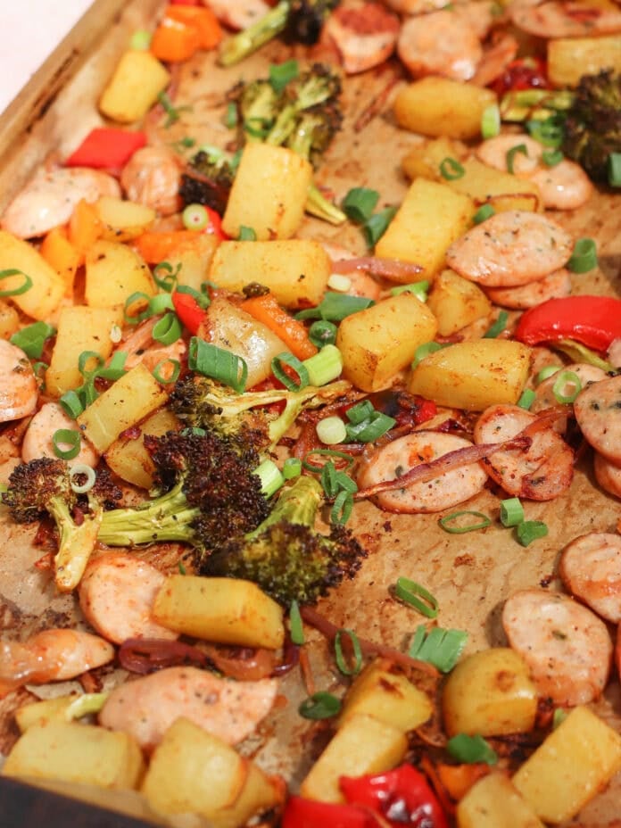 A sheet pan filled with roasted sausage slices, pineapple chunks, broccoli florets, red and yellow bell peppers, and chopped green onions. The ingredients are lightly browned and seasoned.