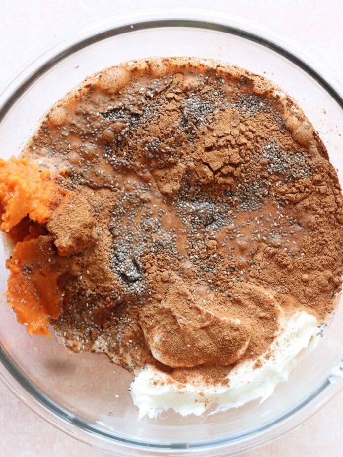 A glass bowl containing pumpkin puree, cream cheese, chia seeds, cinnamon, and cocoa powder, before mixing, on a light pink surface.