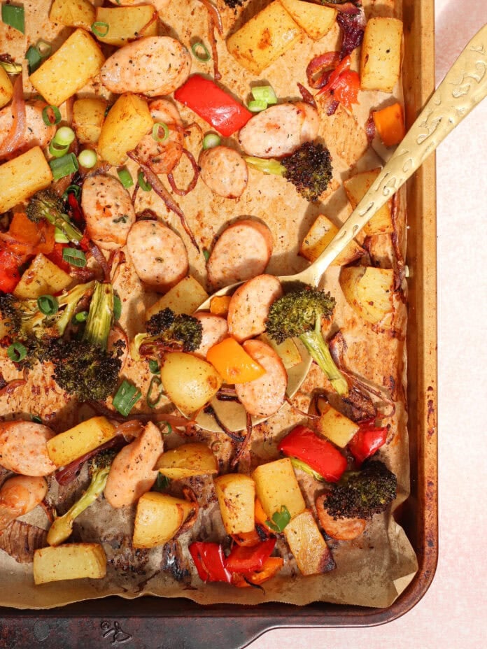 A sheet pan filled with roasted sausage slices, diced potatoes, broccoli, red and yellow bell peppers, and onions, garnished with chopped green onions, with a serving spoon on the right side.
