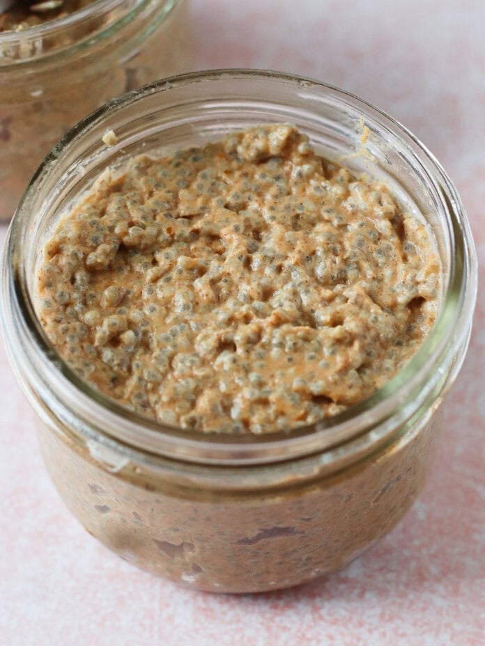 A small glass jar filled with a thick, textured mixture that appears to be a type of overnight oats or chia pudding, placed on a light pink surface. Another partially visible jar is in the background.