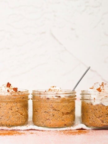Three small glass jars filled with pumpkin chia pudding, topped with whipped cream and sprinkled with cinnamon. A spoon is placed in the middle jar. The jars are set on a lightly dusted surface against a white textured wall.