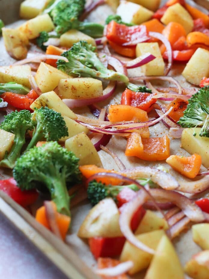 A baking sheet lined with parchment paper holds chopped vegetables including broccoli, red and orange bell peppers, red onion, and potatoes, all seasoned and ready to be roasted.