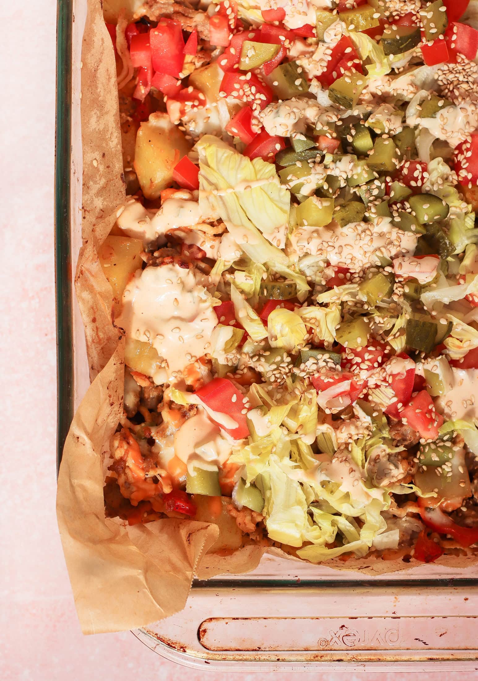 A close-up of a Big Mac casserole in a glass dish, topped with chopped lettuce, diced tomatoes, pickles, sesame seeds, and a drizzle of creamy sauce. The dish is lined with parchment paper.