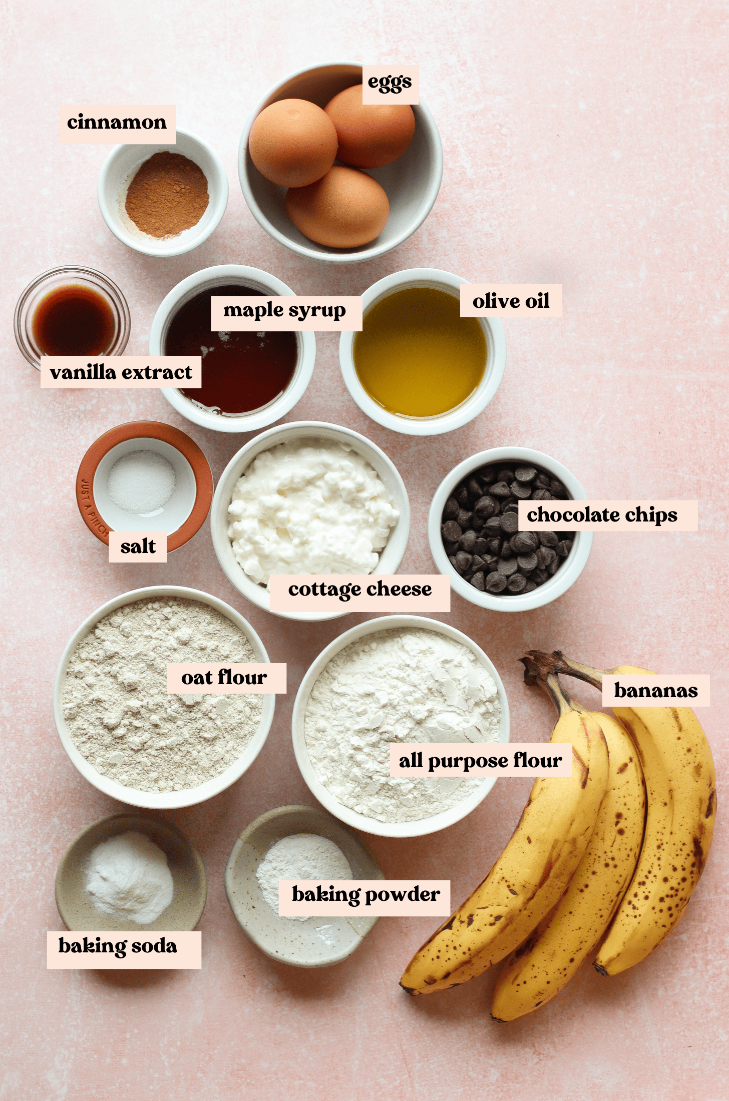 A flat lay of baking ingredients on a pink surface, perfect for cottage cheese banana bread, including eggs, cinnamon, vanilla extract, maple syrup, olive oil, salt, chocolate chips, oat flour, bananas, and more.