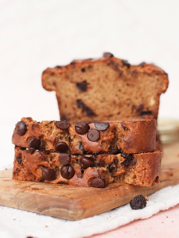 Two slices of chocolate chip banana bread rest on a wooden board, with the remaining loaf in the background. The bread is golden brown and studded with melted chocolate chips.