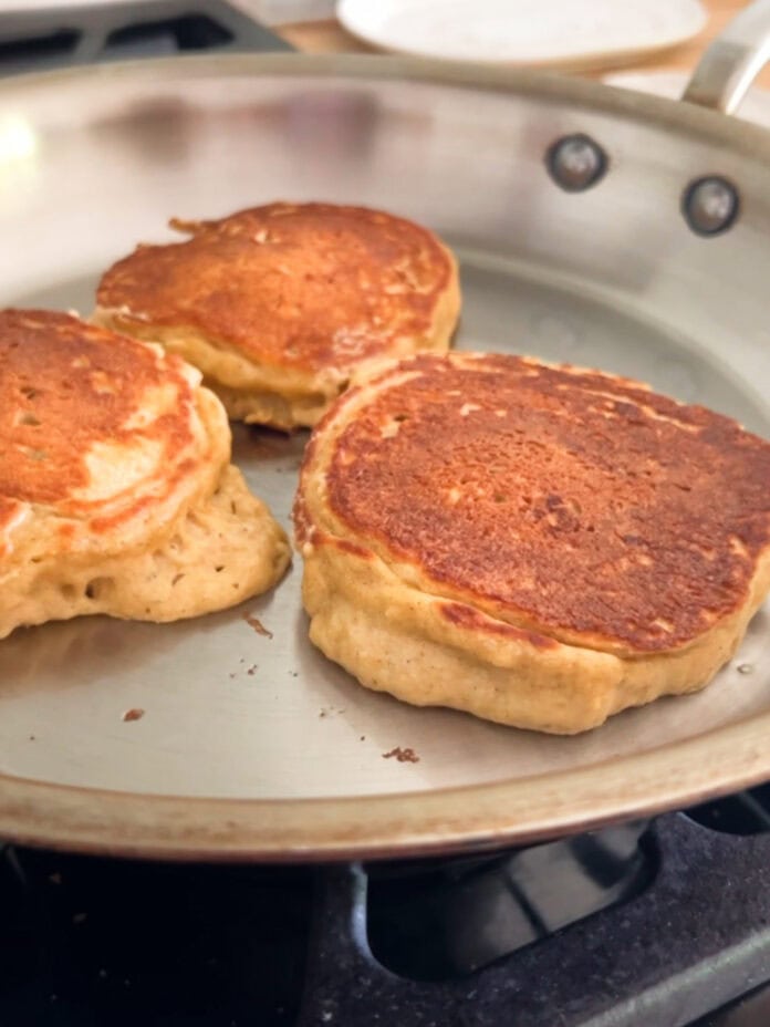 Three thick, golden-brown Greek yogurt pancakes are cooking in a stainless steel skillet on a stovetop. The pancakes appear fluffy and slightly crisp on the outside.