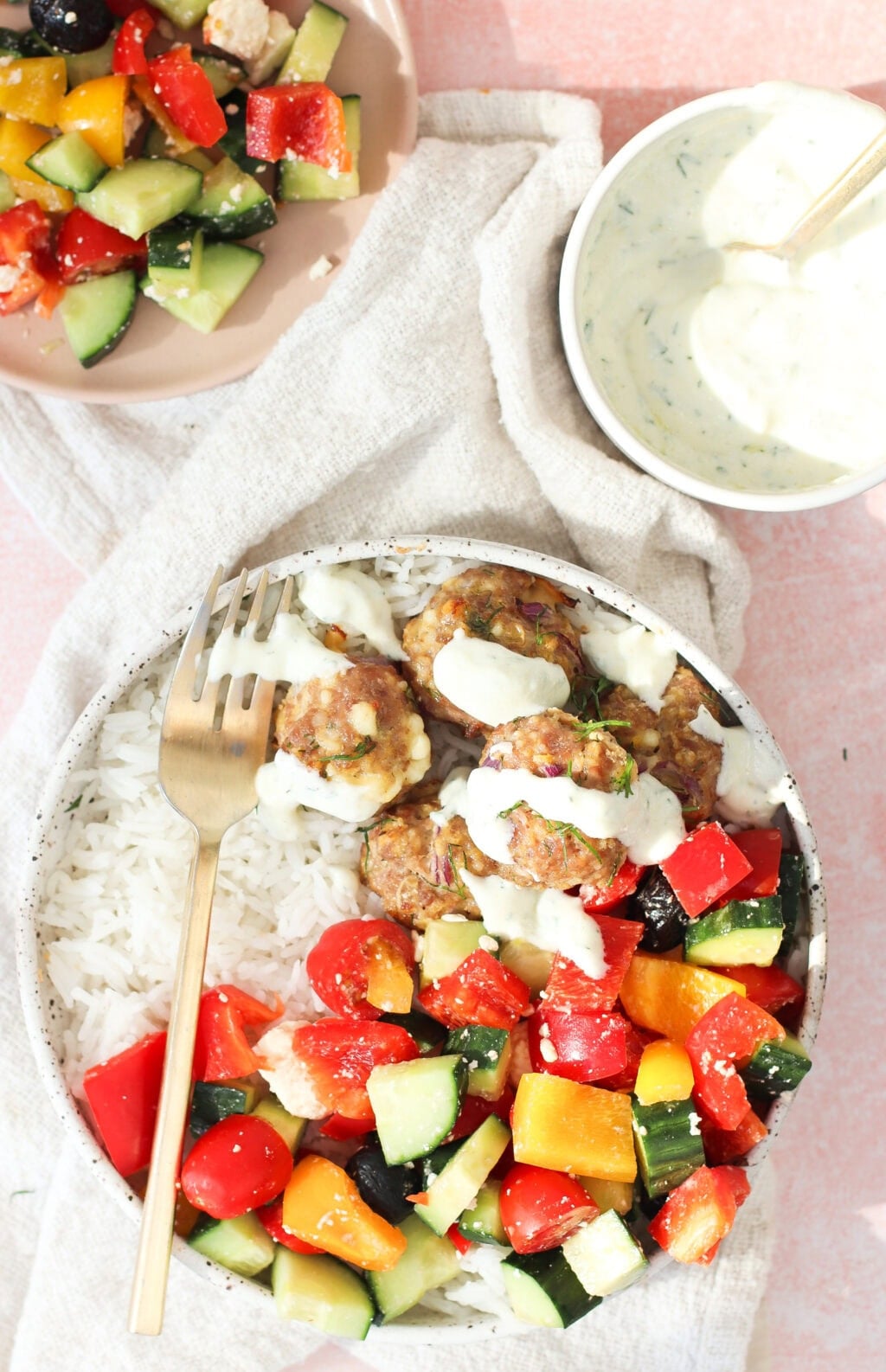 A bowl of white rice topped with colorful chopped vegetables, meatballs, and creamy sauce, with a fork resting on the bowl. Nearby are a small plate of salad and a bowl of sauce with a spoon on a light cloth.
