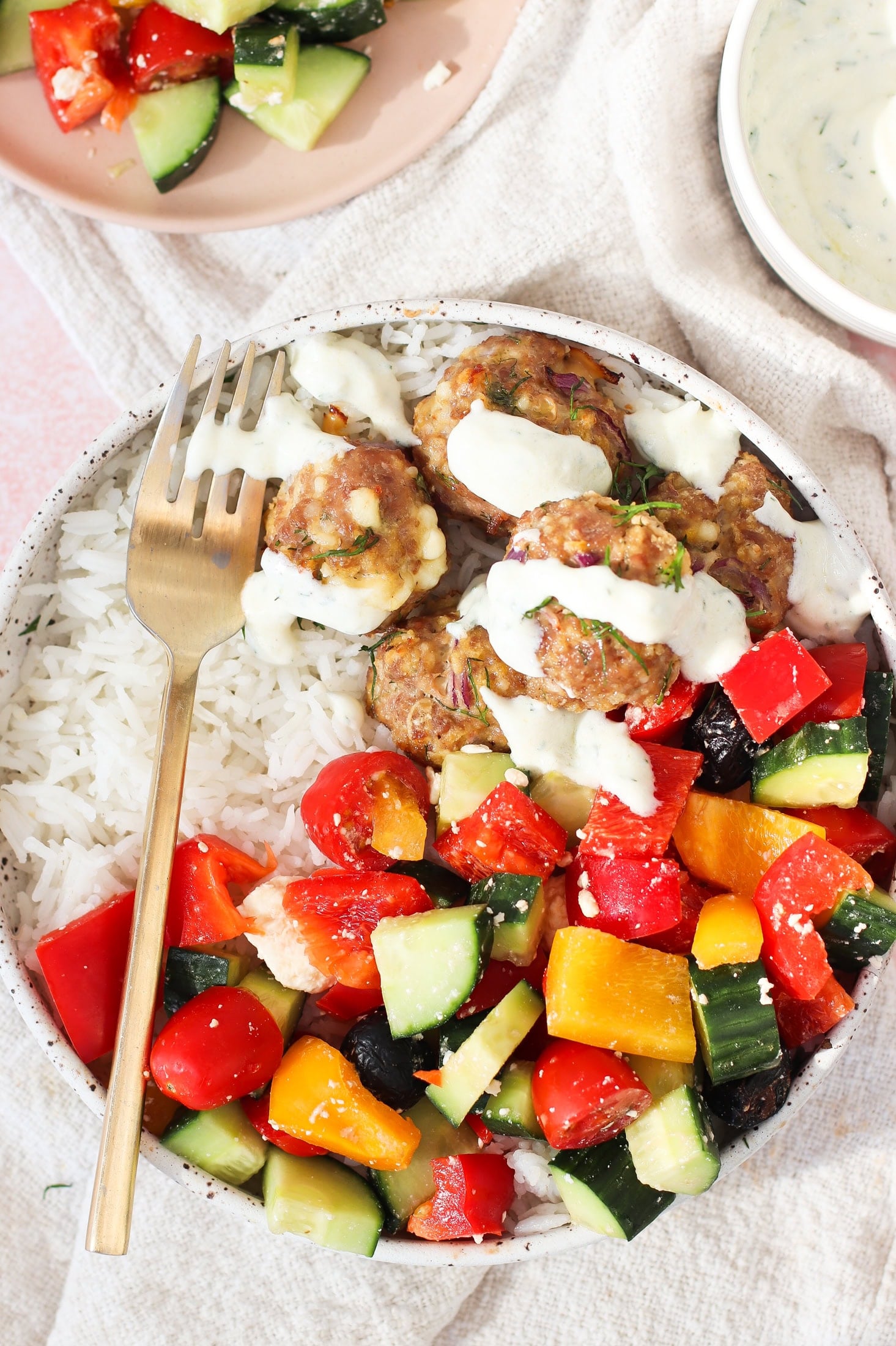 A bowl of white rice topped with meatballs drizzled with white sauce, served with a colorful salad of cucumbers, tomatoes, bell peppers, and olives; a fork rests on the bowl.