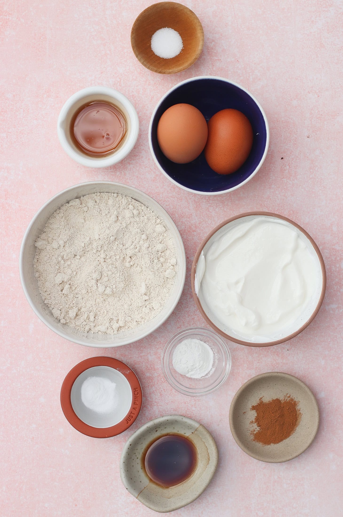 A flat lay of baking ingredients on a pink surface: two eggs, a bowl of oat flour, yogurt, ground cinnamon, baking powder, vanilla extract, baking soda, maple syrup, and a pinch of salt in small bowls.