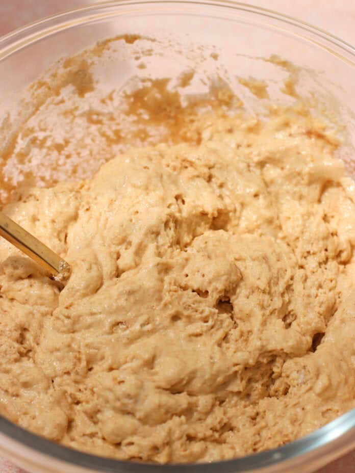 A close-up of thick, sticky bread dough being mixed in a clear glass bowl with a spoon, set on a light pink surface.