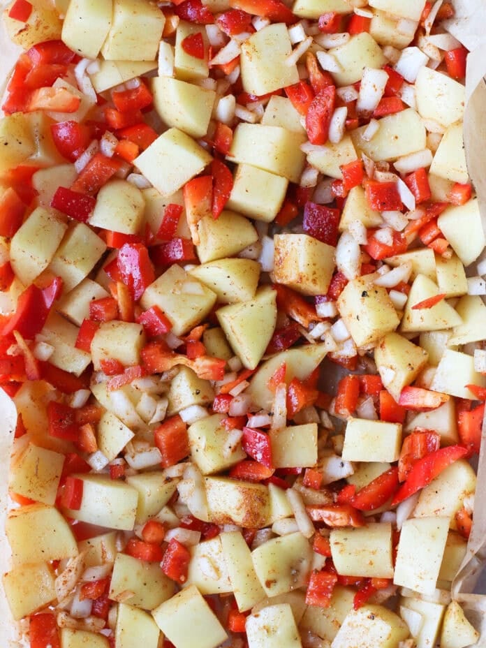 Diced potatoes, red bell peppers, and chopped onions are spread out on a parchment-lined baking sheet, seasoned and ready for roasting.
