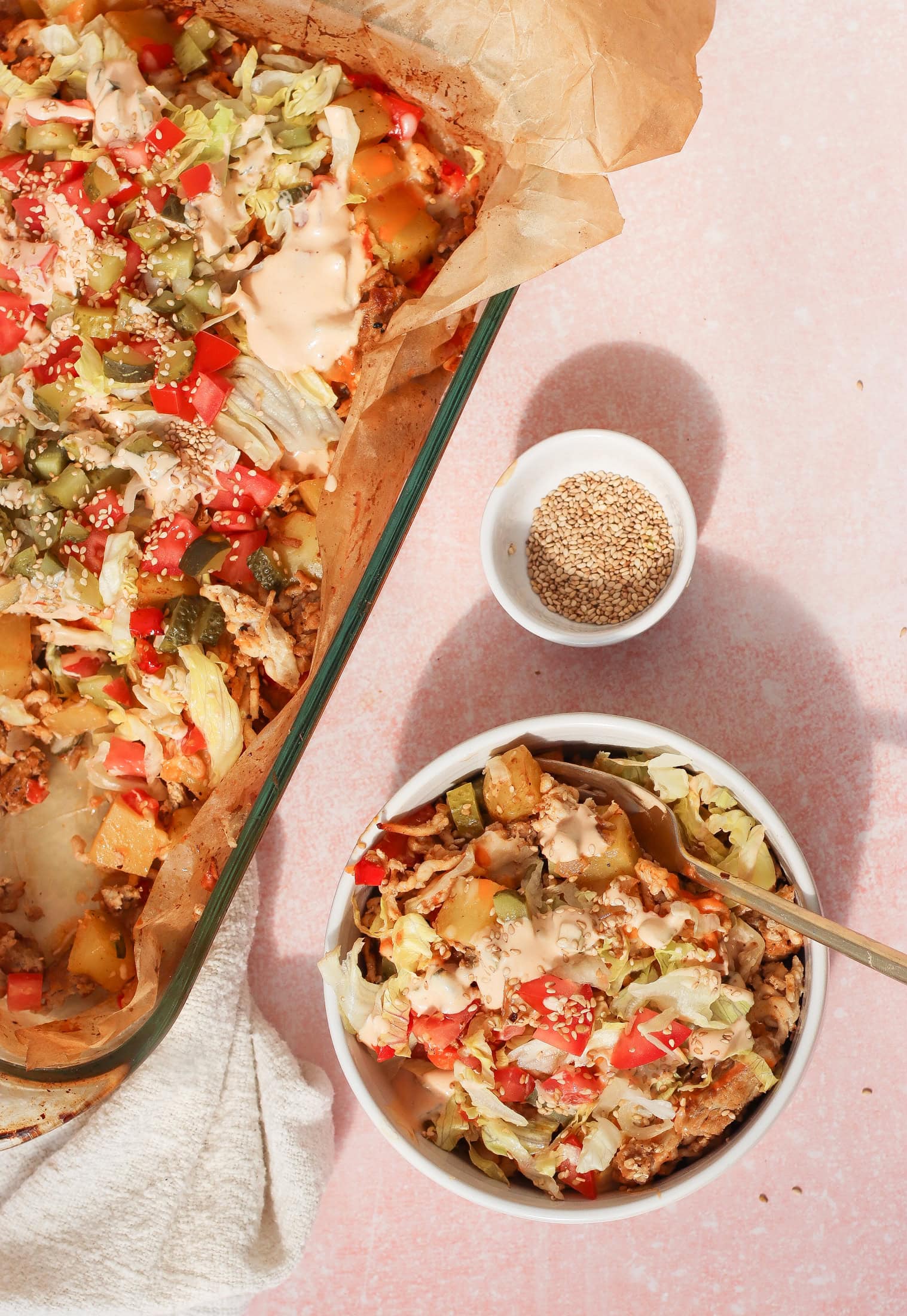 A bowl and baking dish filled with a layered casserole of shredded lettuce, diced tomatoes, pickles, potatoes, and creamy sauce, topped with sesame seeds. A small bowl of sesame seeds sits nearby on a pink surface.