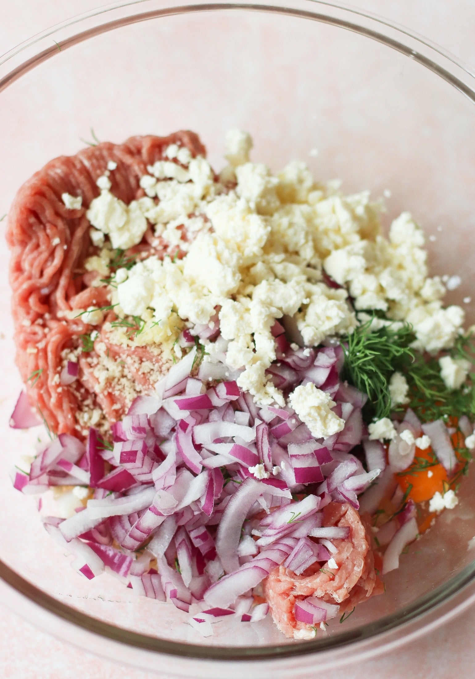 A glass bowl containing ground meat, crumbled feta cheese, chopped red onion, fresh dill, a cracked egg, and breadcrumbs, ready to be mixed for a recipe.