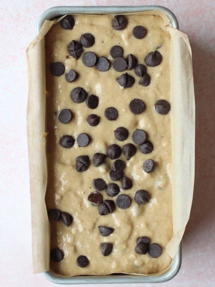 A loaf pan lined with parchment paper is filled with banana bread batter, topped with scattered chocolate chips, ready to be baked.