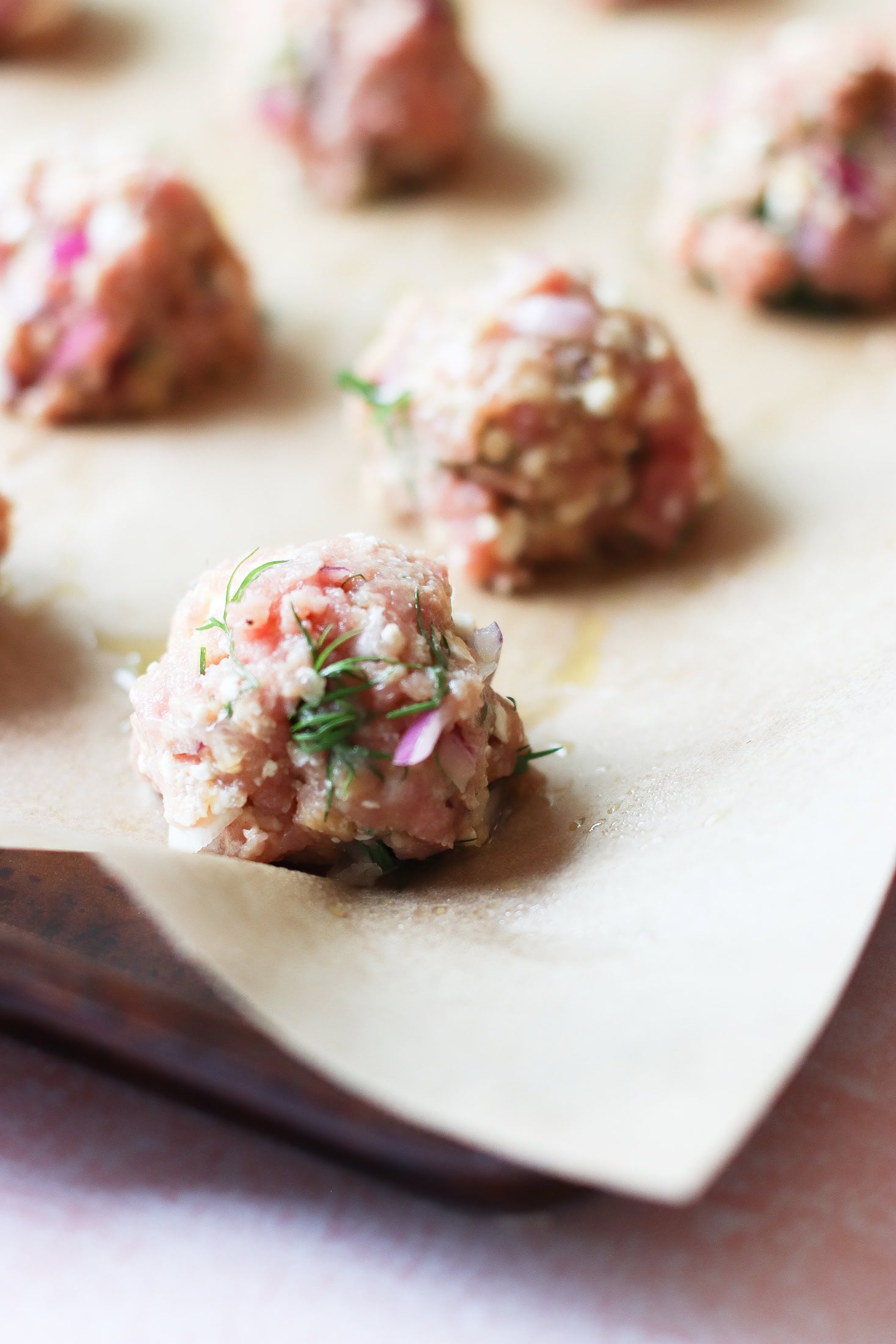 A close-up of raw meatballs with visible herbs and red onion pieces, resting on parchment paper on a baking tray.