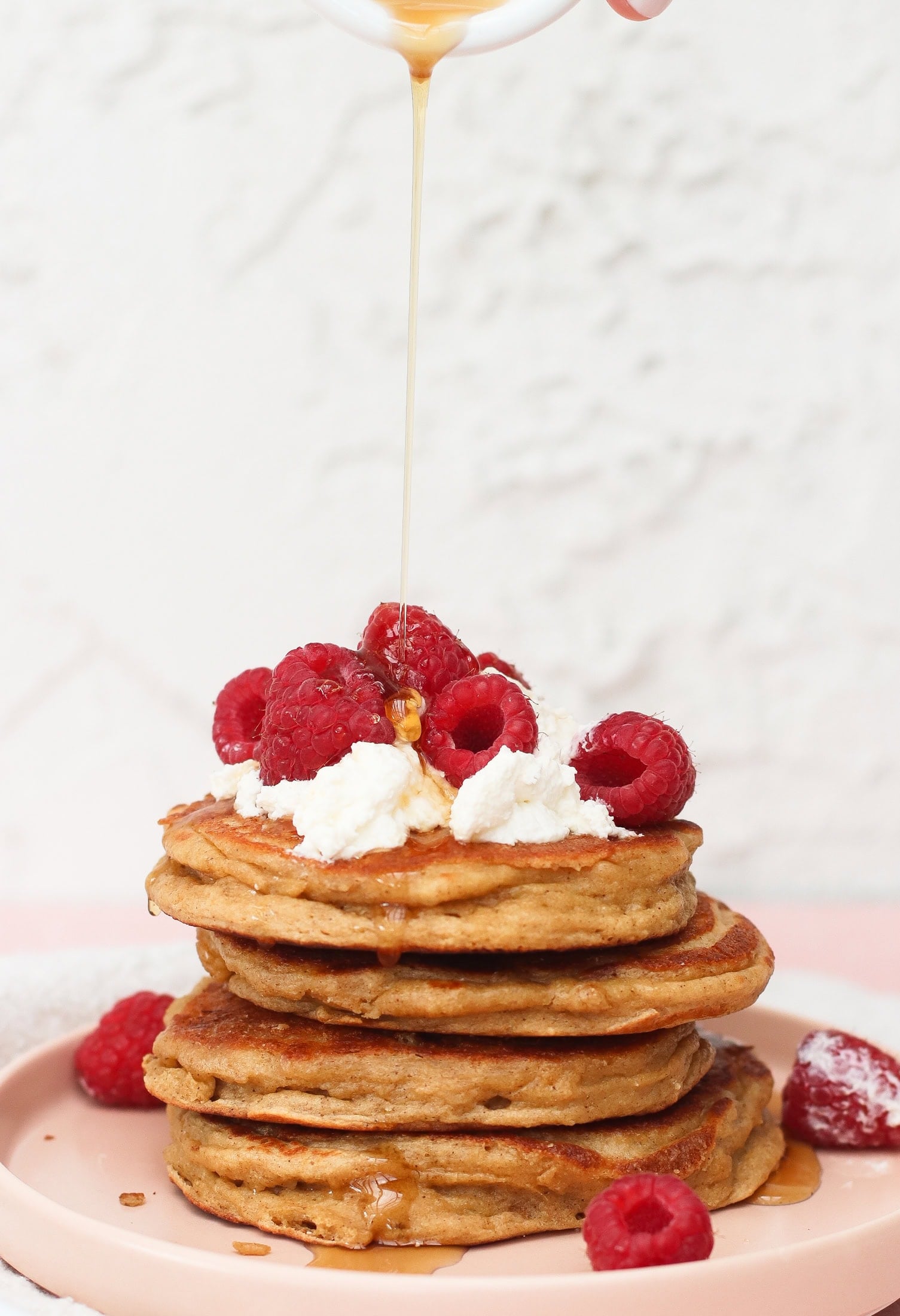 A stack of pancakes topped with whipped cream and fresh raspberries is being drizzled with syrup. More raspberries are scattered on the pink plate below.