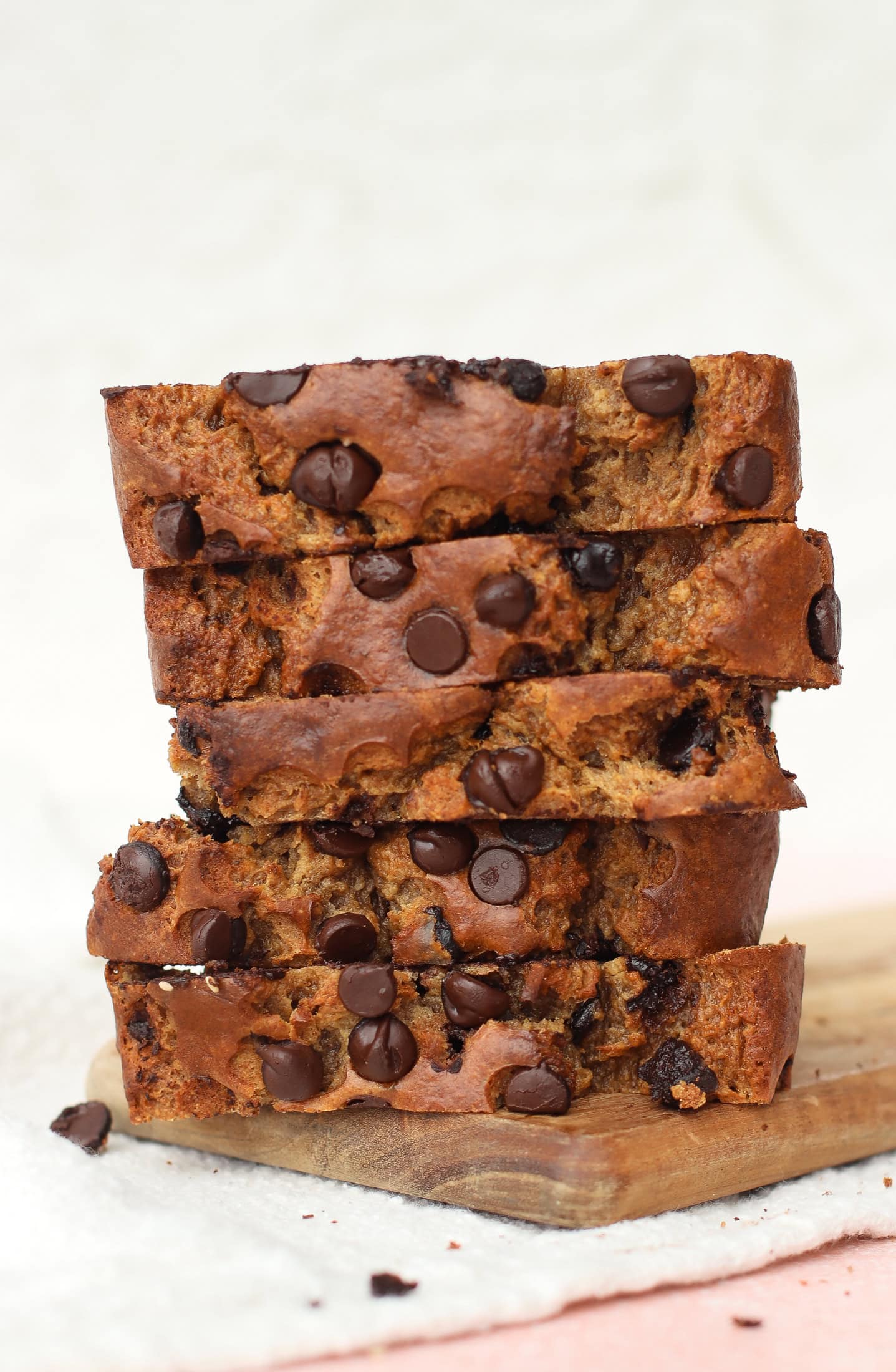 A stack of five thick slices of chocolate chip cottage cheese banana bread sits on a wooden board, with chocolate chips visible throughout the moist bread. The background is light and slightly blurred.