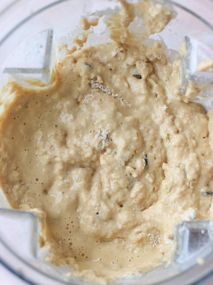 A close-up view of thick, lumpy batter with visible chunks, inside a clear blender container. The mixture appears creamy and lightly golden in color.