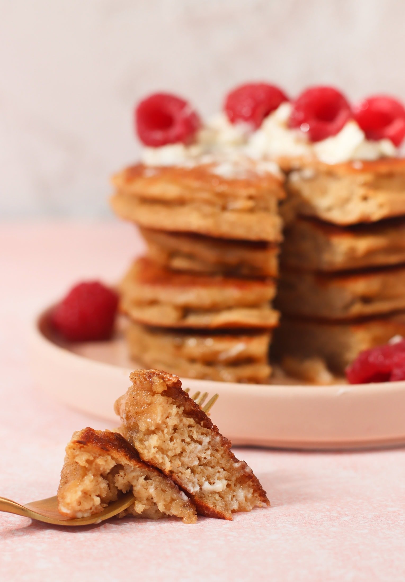 A close-up of a stack of Greek yogurt pancakes topped with raspberries and melting whipped cream on pink a plate, with a fork holding a bite-sized piece in the foreground. The background is soft pink and slightly blurred.