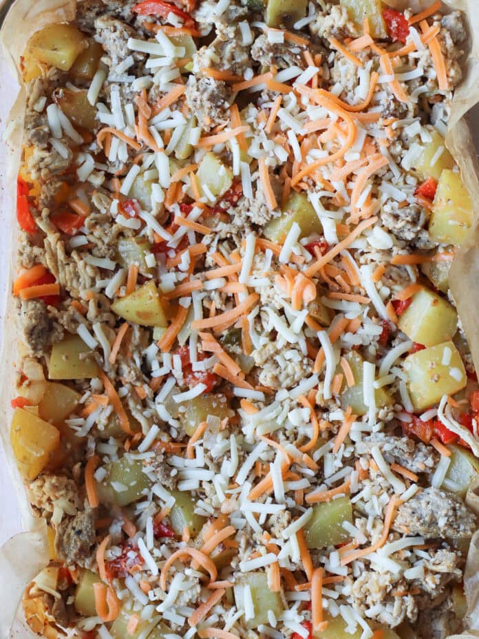 A close-up of a casserole dish filled with diced potatoes, red bell peppers, sausage, and shredded cheese on top, all on parchment paper and ready for baking.