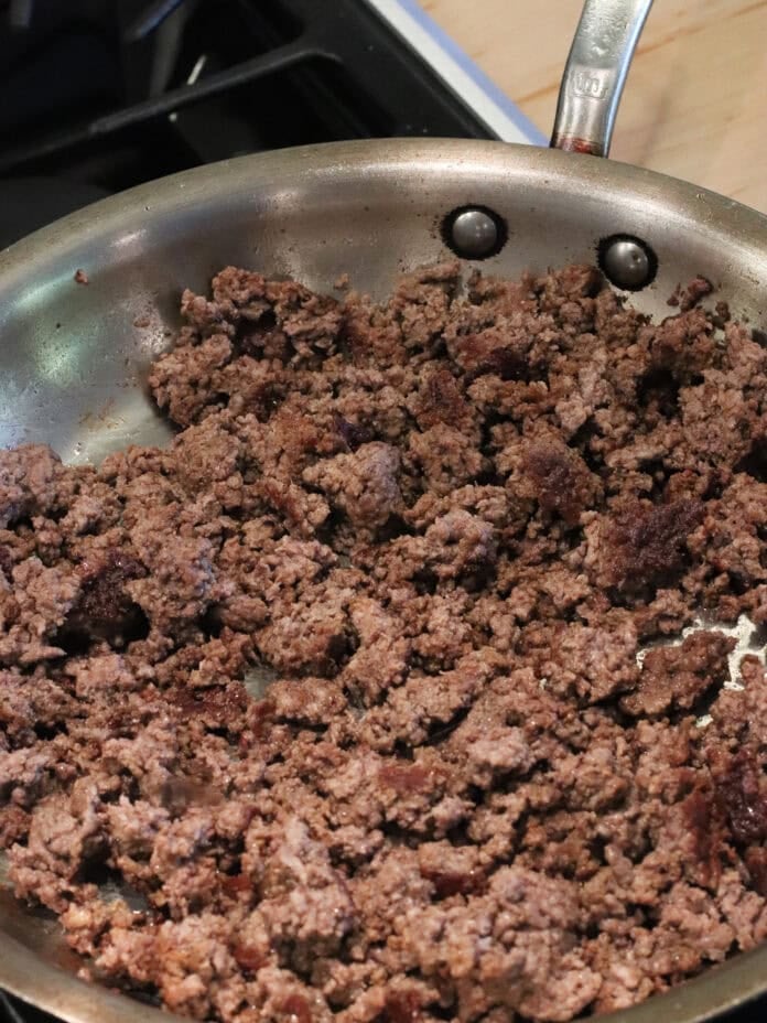 Cooked ground beef in a stainless steel skillet on a stove, with browned and crumbled meat pieces visible.