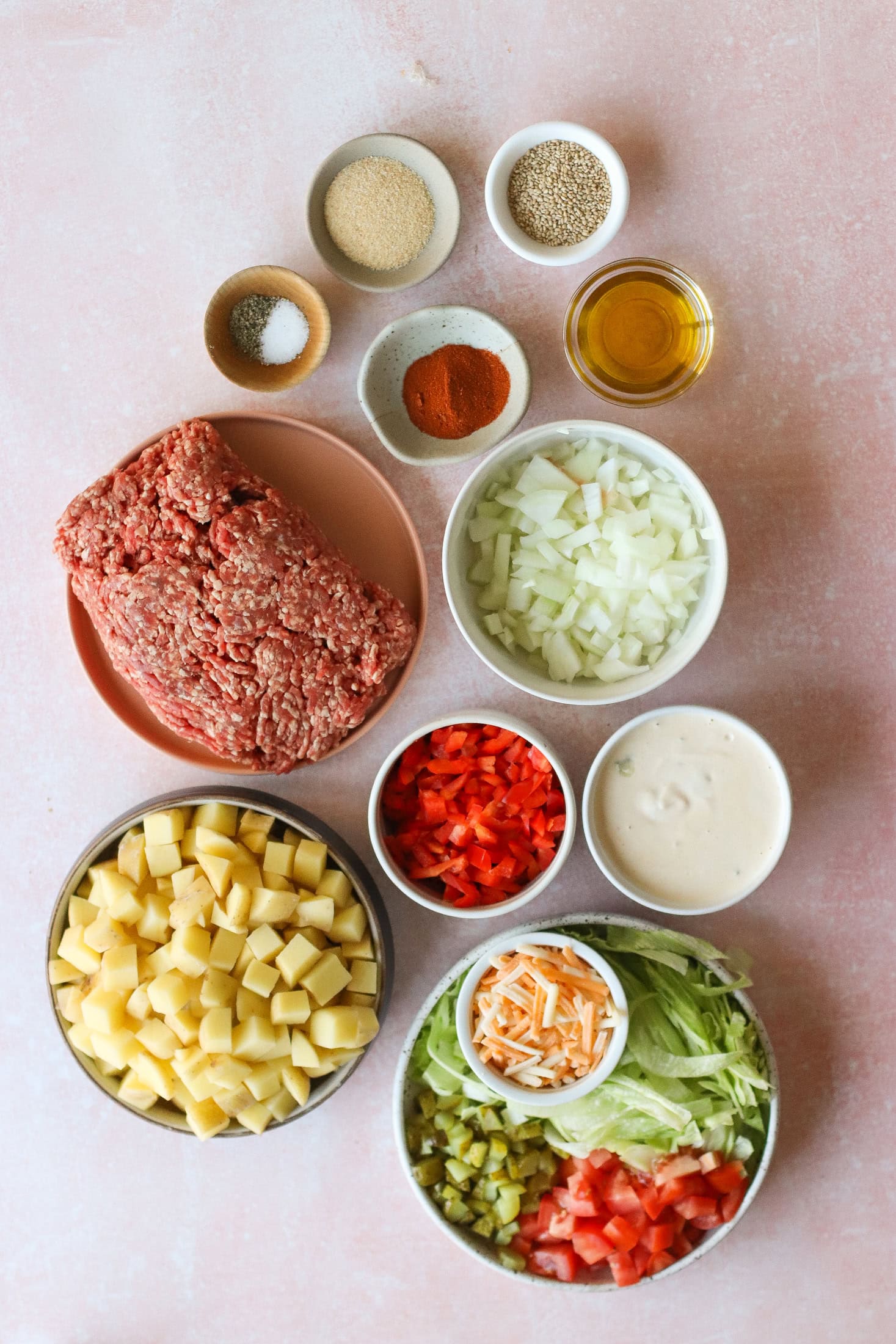 Overhead view of ingredients in small bowls on a pink surface, including ground beef, diced potatoes, chopped onions, diced red pepper, shredded lettuce, diced tomatoes, chopped pickles, shredded cheese, creamy sauce, spices, and oil.