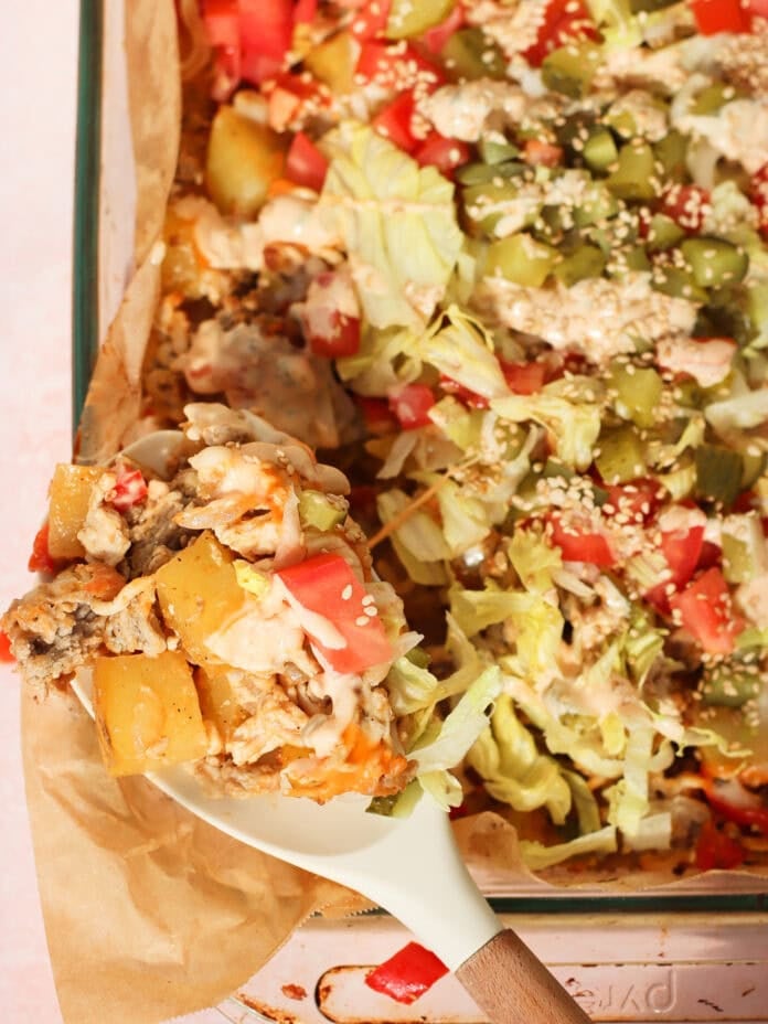 A close-up of a casserole dish filled with a layered baked meal including diced potatoes, chopped lettuce, tomatoes, pickles, sesame seeds, and a creamy sauce, with a serving spoon lifting out a portion.