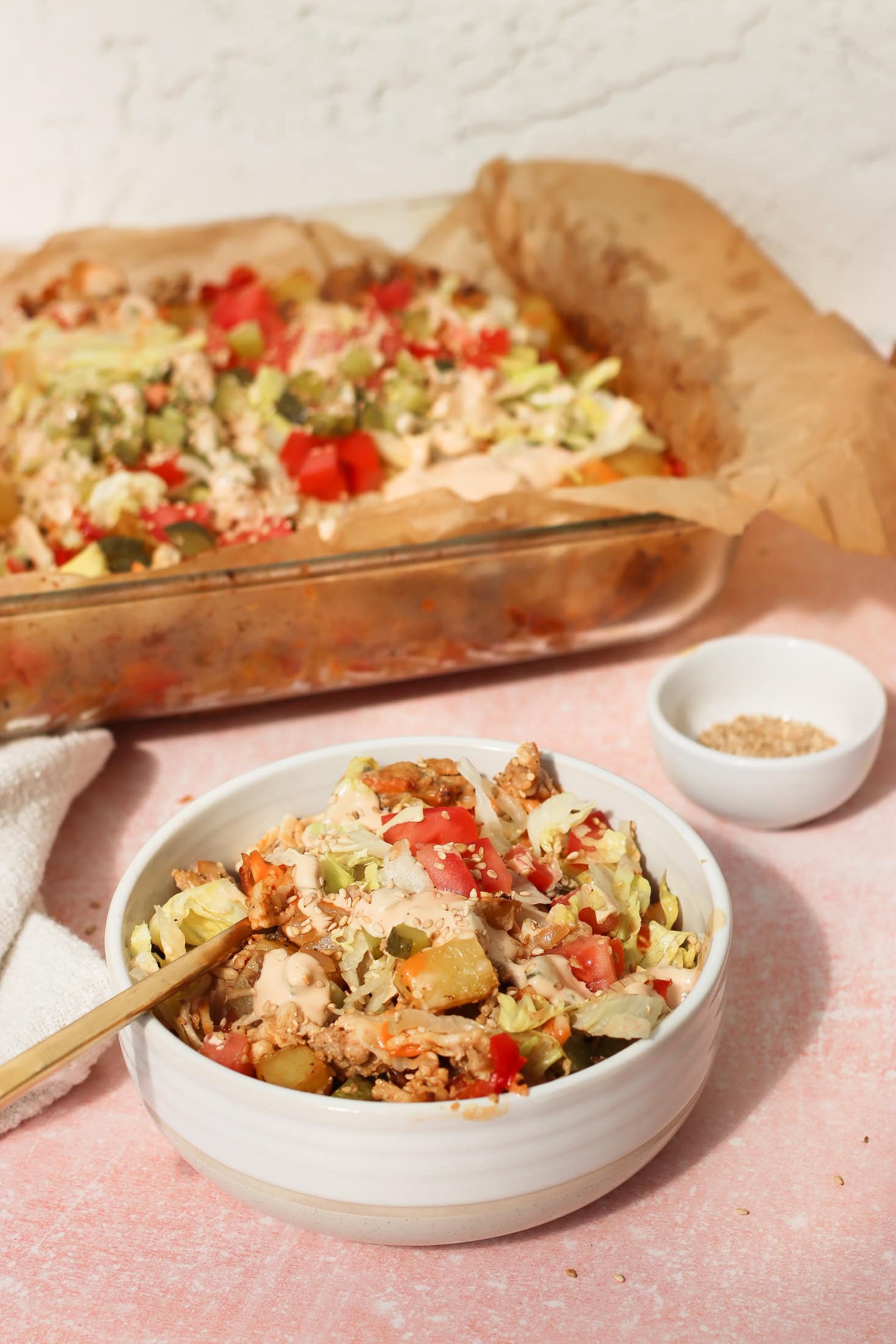 A white bowl filled with a mixture of vegetables, ground meat, and sauce sits in front of a glass baking dish with the same casserole. A spoon is in the bowl, and a small dish of seasoning is nearby on a pink surface.