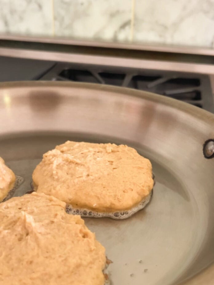 Three uncooked pancakes are cooking in a stainless steel skillet on a stovetop, with bubbles forming around the edges of the dough