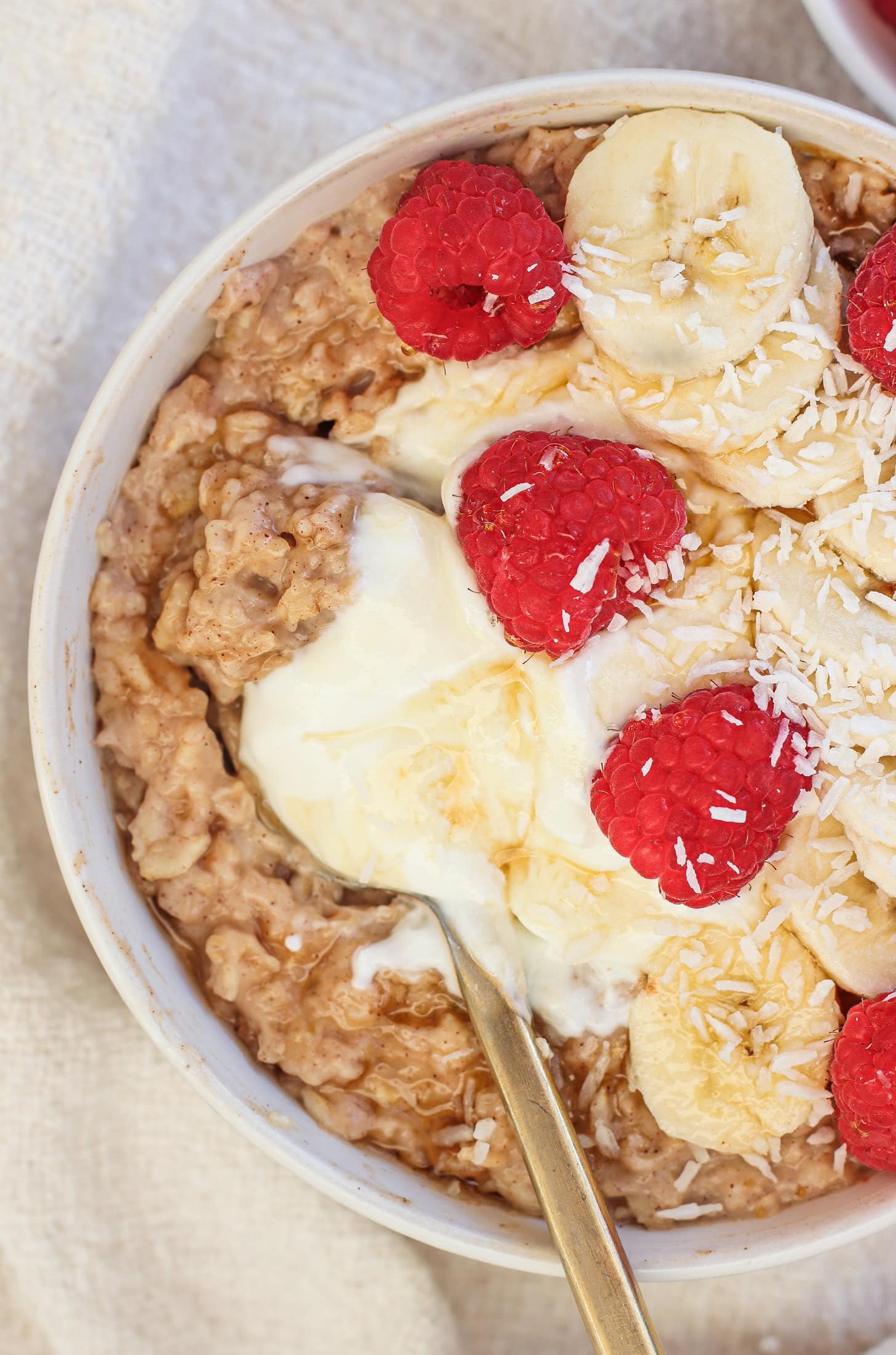 A bowl of high protein oatmeal topped with banana slices, raspberries, shredded coconut, and a dollop of yogurt. A spoon rests in the bowl, set on a light-colored fabric.