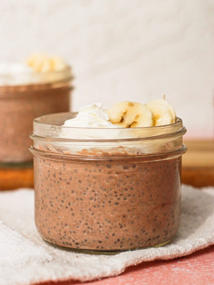 A small glass jar filled with chocolate chia pudding, topped with whipped cream and banana slices, sits on a white cloth. Another similar jar is blurred in the background.