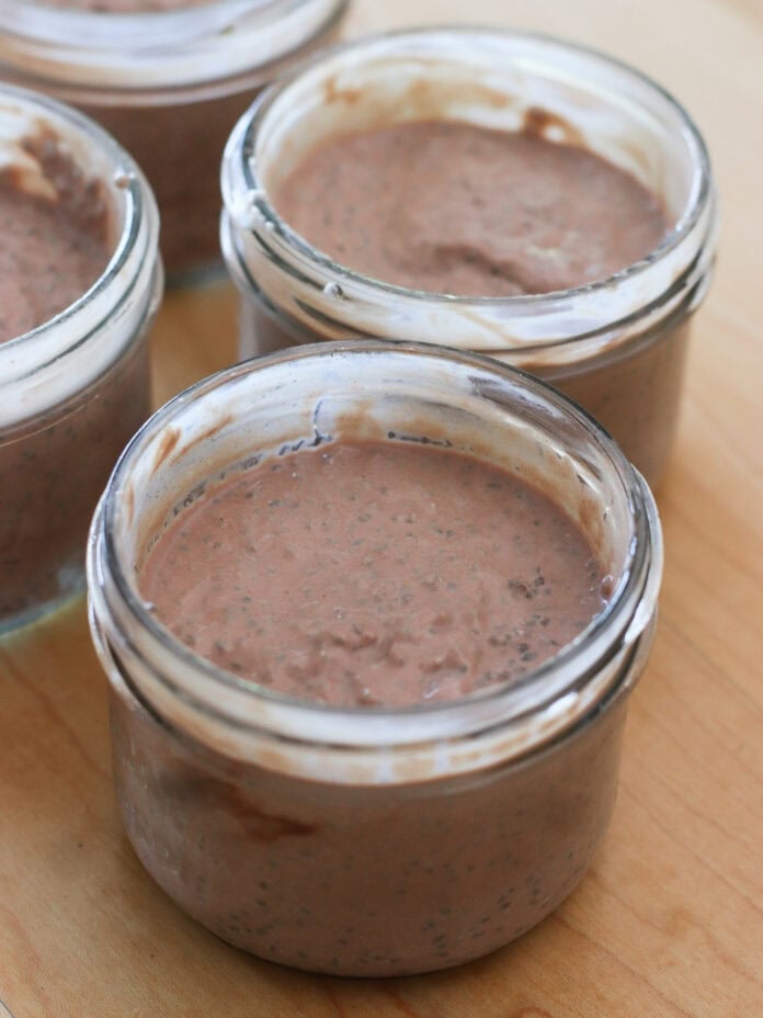 Four small glass jars filled with chocolate chia pudding are placed on a wooden surface. The pudding is brown and has a creamy texture with visible chia seeds.