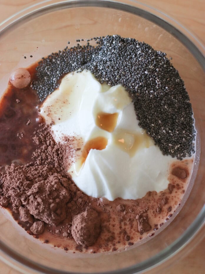 A glass bowl containing yogurt, cocoa powder, chia seeds, vanilla extract, and milk, all unmixed, on a wooden surface.