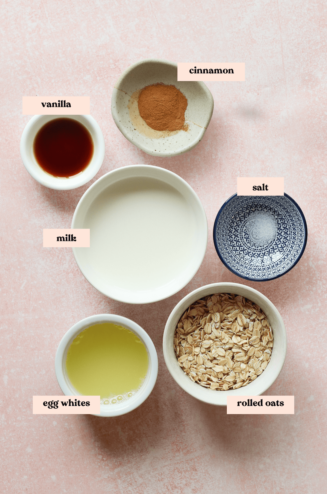 Six bowls on a pink surface contain labeled ingredients for high protein oatmeal: vanilla, milk, egg whites, cinnamon, salt, and rolled oats, all neatly arranged for recipe preparation.