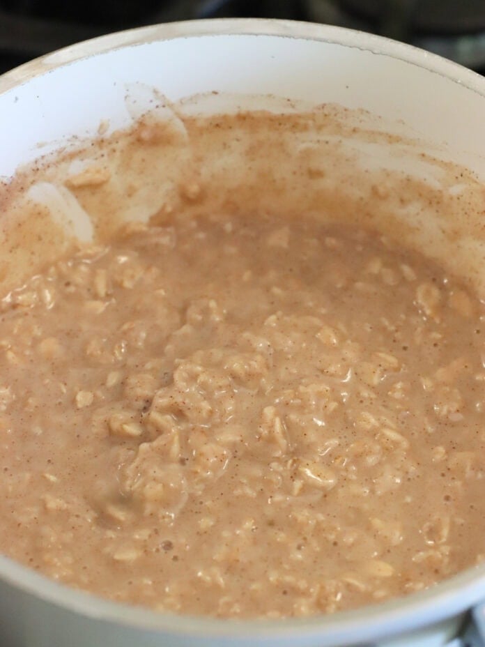 A close-up of a pot filled with creamy, high protein oatmeal, lightly brown in color, being cooked on a stove. The oats appear well mixed with most of the liquid absorbed