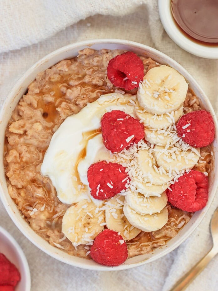 A bowl of high protein oatmeal topped with sliced bananas, raspberries, shredded coconut, a dollop of yogurt, and a drizzle of syrup, placed on a light-colored cloth next to a small bowl of syrup and a spoon.