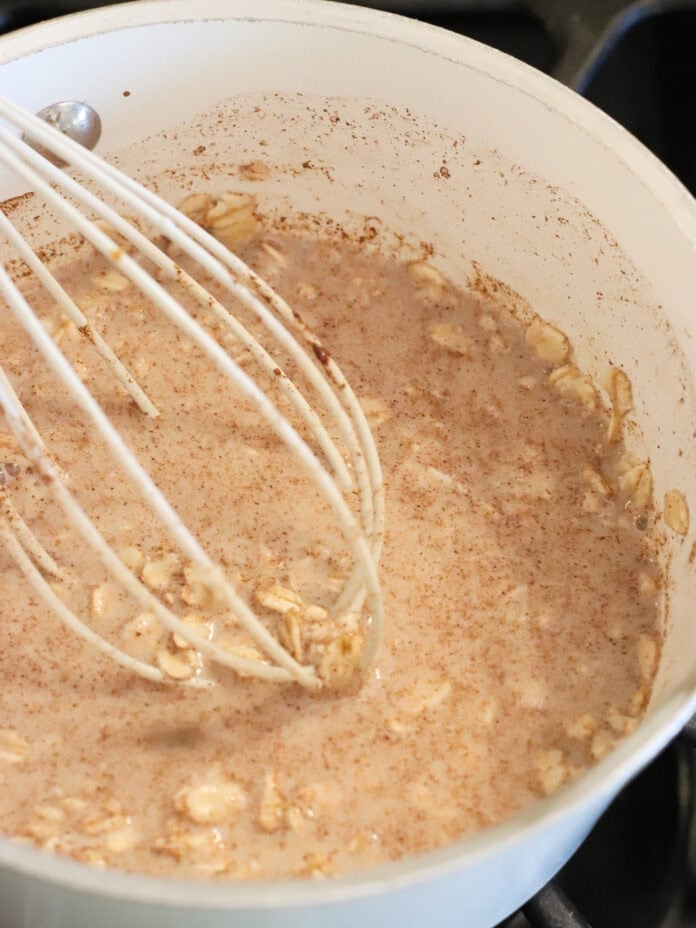 A white saucepan on a stove contains oatmeal being mixed with a white whisk. The mixture appears creamy with visible oats and a sprinkle of cinnamon or spice on top.