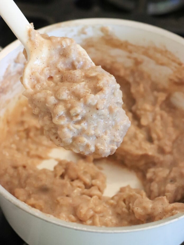 A close-up of a white spoon lifting a creamy, thick oatmeal mixture with visible specks of cinnamon from a white pot. The background is slightly blurred.