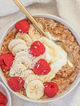A bowl of oatmeal topped with banana slices, raspberries, shredded coconut, and a dollop of yogurt, with a gold spoon resting inside. A small dish of raspberries is visible in the corner.