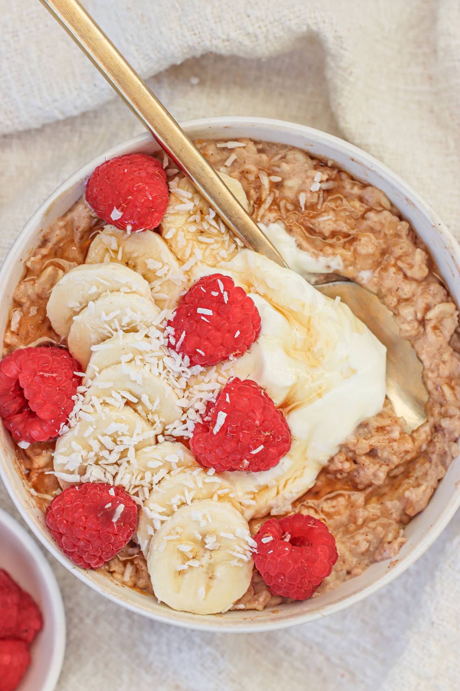 A bowl of oatmeal topped with banana slices, raspberries, shredded coconut, and a dollop of yogurt, with a gold spoon resting inside. A small dish of raspberries is visible in the corner.