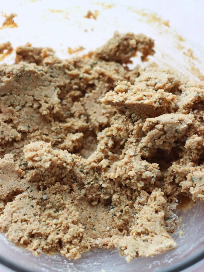 A close-up of cookie dough in a glass mixing bowl, showing a coarse and crumbly texture with visible flecks of ingredients throughout.