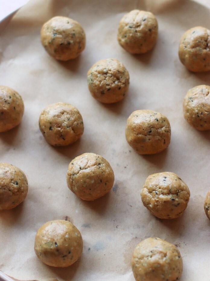Sixteen round dough balls evenly spaced on a baking sheet lined with parchment paper, ready to be baked. The dough appears to have herbs or seasoning mixed in.