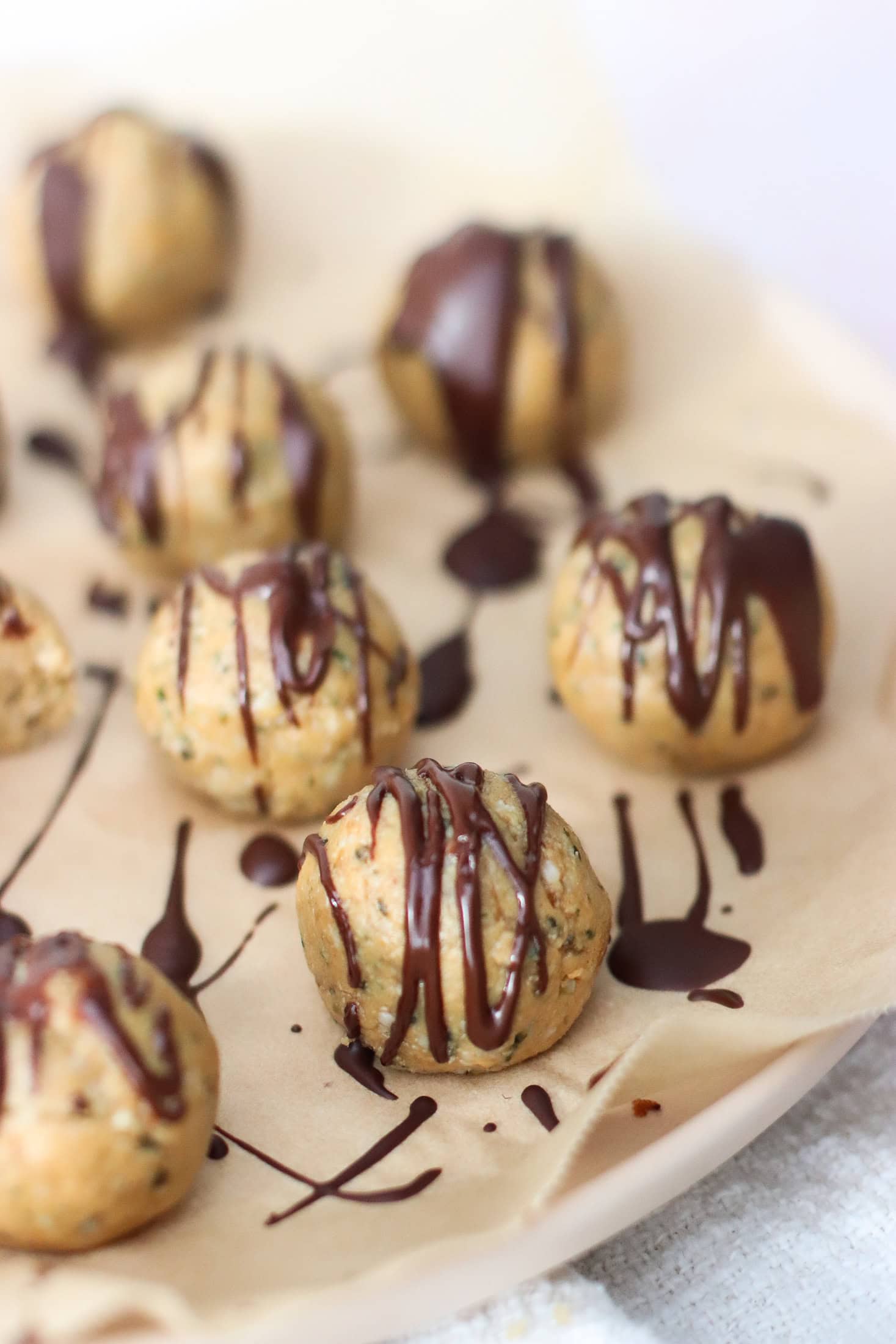 Round, bite-sized cookie balls on parchment paper are drizzled with dark chocolate. The treats appear homemade, with a nutty, textured surface and a glossy chocolate topping. Some are in focus, while others are blurred in the background.