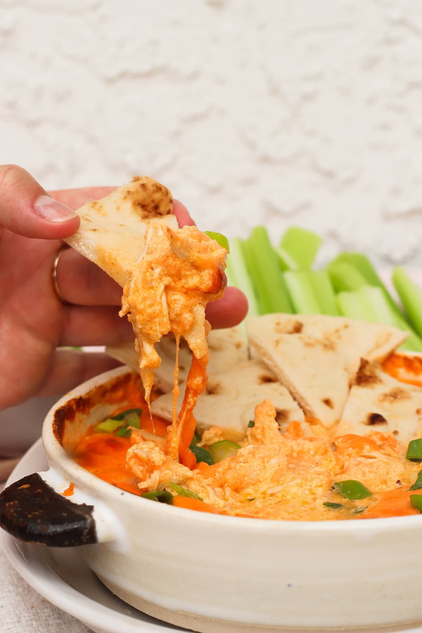 A hand dips a piece of pita bread into a cheesy, orange buffalo chicken dip in a white dish. Celery sticks are visible in the background.