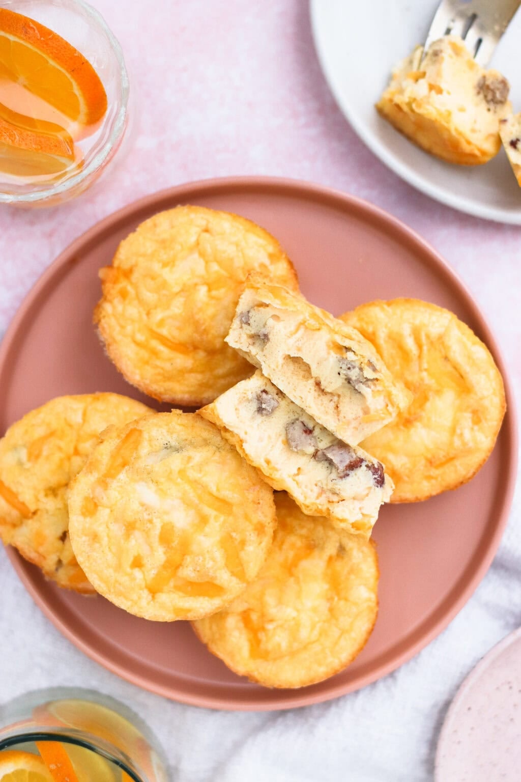A pink plate holds several mini egg muffins, with one muffin cut in half to show a filling of cheese and pieces of sausage. A glass of orange drink and a plate with another muffin piece are nearby.