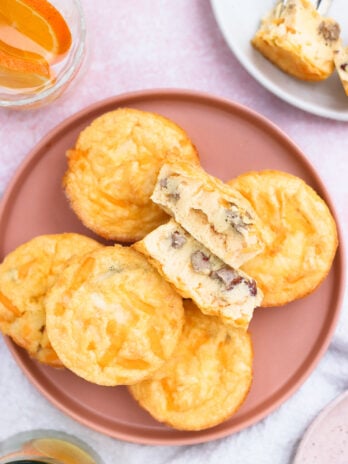 A pink plate holds several mini egg muffins, with one muffin cut in half to show a filling of cheese and pieces of sausage. A glass of orange drink and a plate with another muffin piece are nearby.