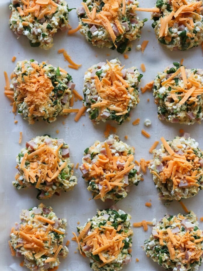 Twelve unbaked patties made of chopped vegetables, ham, and cheese are arranged on a parchment-lined baking tray, each topped with a small mound of shredded cheddar cheese.